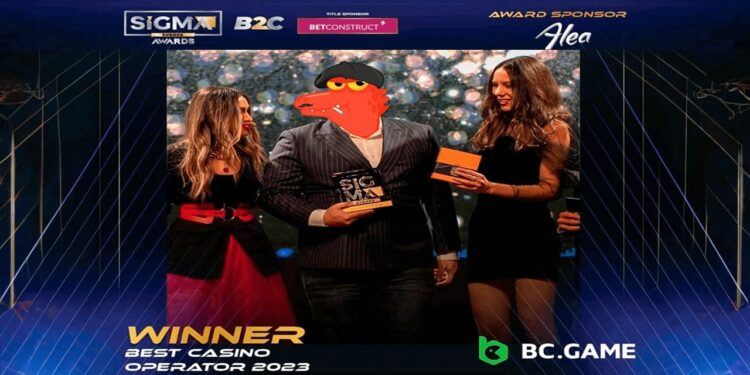 Dos mujeres y un hombre con cara de cocodrilo sosteniendo un premio sobre el escenario.