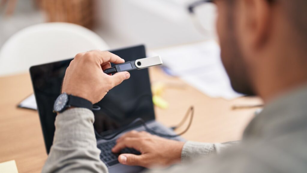 Una persona sostiene una hardware wallet frente a una laptop