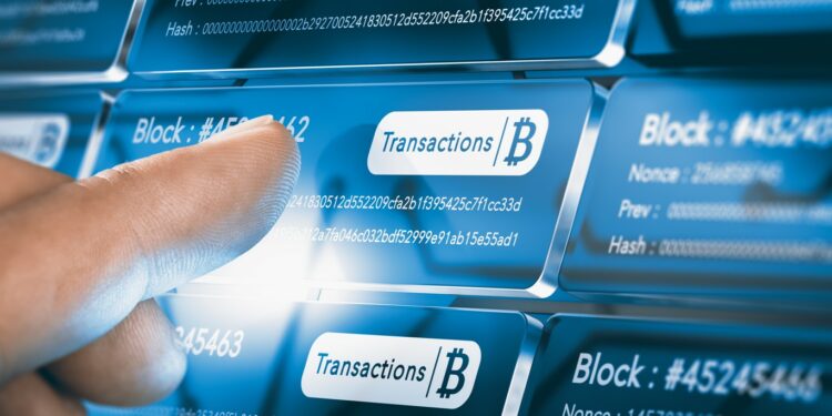 Una mano a punto de presionar una pantalla donde se lee la palabra transacción con símbolos de bitcoin
