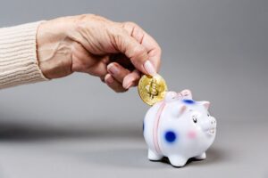 La mano de una persona introduciendo una moneda física con el símbolo dorado de Bitcoin dentro de la ranura de una alcancía tradicional de cerámica con forma de cerdito. El fondo es limpio poniendo todo el énfasis visual en el contraste entre el método de ahorro clásico y el activo digital.