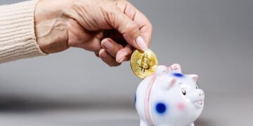 La mano de una persona introduciendo una moneda física con el símbolo dorado de Bitcoin dentro de la ranura de una alcancía tradicional de cerámica con forma de cerdito. El fondo es limpio poniendo todo el énfasis visual en el contraste entre el método de ahorro clásico y el activo digital.