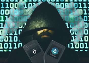 Una persona con el rosto cubierto, wallets de trezor y código al fondo