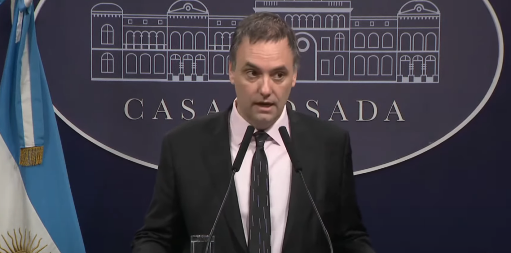 Fotografía de plano medio de Guillermo Adorni, funcionario público argentino, vestido con traje formal oscuro y camisa clara. Se encuentra hablando frente a un micrófono en un atril de prensa oficial, con un fondo institucional neutro y desenfocado.
