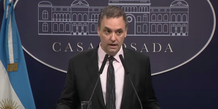 Fotografía de plano medio de Guillermo Adorni, funcionario público argentino, vestido con traje formal oscuro y camisa clara. Se encuentra hablando frente a un micrófono en un atril de prensa oficial, con un fondo institucional neutro y desenfocado.