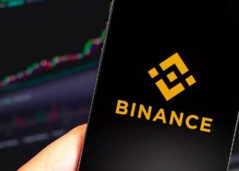 Teléfono móvil con logo de Binance en la pantalla y un gráfico de velas al fondo en un ordenador.