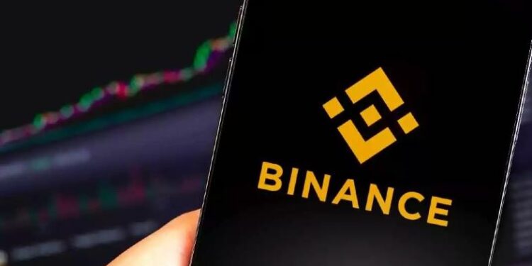 Teléfono móvil con logo de Binance en la pantalla y un gráfico de velas al fondo en un ordenador.