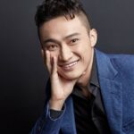 Justin Sun sonriendo delante de un fondo negro apoyando su mano en la cara.