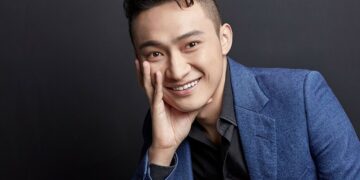 Justin Sun sonriendo delante de un fondo negro apoyando su mano en la cara.