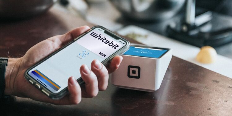 Una mano varonil sostiene un teléfono celular con la app de WhiteBIT abierta cerca de de un punto de venta.