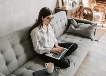 Mujer joven sentada en un sofá gris con una laptop sobre sus piernas.