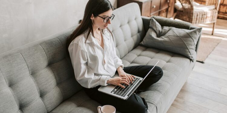 Mujer joven sentada en un sofá gris con una laptop sobre sus piernas.