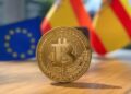 Una imagen con una moneda de bitcoin en primer plano y de fondo una bandera de España y la Unión Europea.