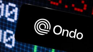 Logo de Ondo Finance en un smarphone con gráficas de precios en el fondo.