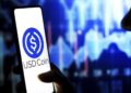 Una persona sostiene un móvil que tiene el logo de USDC en la pantalla, con gráficos de precio al fondo