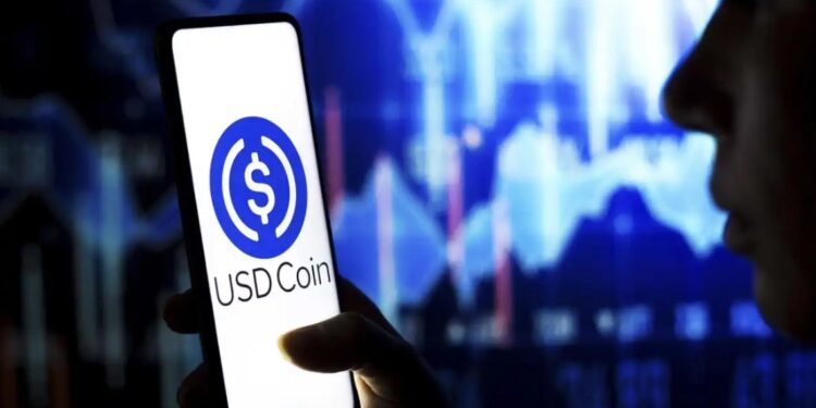 Una persona sostiene un móvil que tiene el logo de USDC en la pantalla, con gráficos de precio al fondo