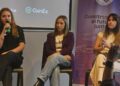 Tres mujeres con ropa casual sentadas conversan con el publico sobre un escenario.