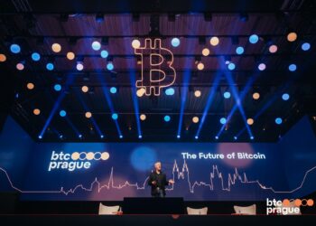 Imagen de un evento o conferencia en un auditorio con un escenario grande. En la pantalla principal del escenario aparece el logo de 'btc prague' y el texto 'The Future of Bitcoin'.