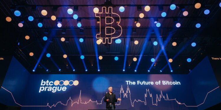 Imagen de un evento o conferencia en un auditorio con un escenario grande. En la pantalla principal del escenario aparece el logo de 'btc prague' y el texto 'The Future of Bitcoin'.