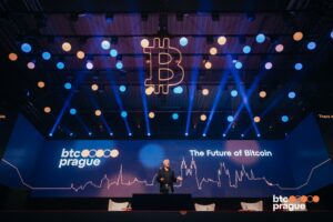 Escenario principal de BTC Prague con un ponente hablando bajo luces azules y naranjas, el logo de Bitcoin colgando y la frase "The Future of Bitcoin" en pantalla.