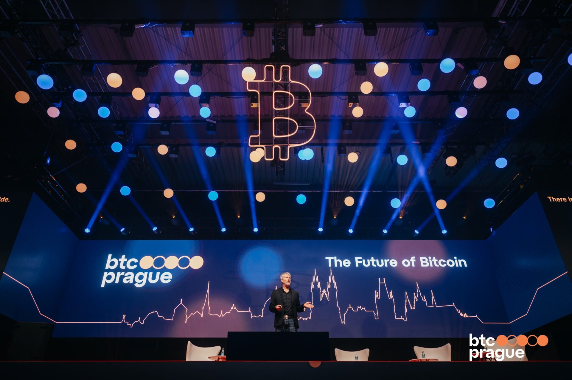 Escenario principal de BTC Prague con un ponente hablando bajo luces azules y naranjas, el logo de Bitcoin colgando y la frase "The Future of Bitcoin" en pantalla.