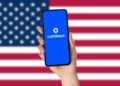 Bandera de Estados Unidos en el fondo. Delante, hay una mano sosteniendo un teléfono en cuya pantalla se lee "Coinbase".