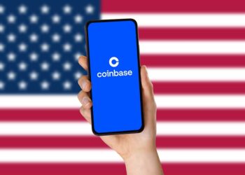 Bandera de Estados Unidos en el fondo. Delante, hay una mano sosteniendo un teléfono en cuya pantalla se lee "Coinbase".
