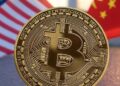 Una moneda dorada con el logo de bitcoin, delante de las banderas de Estados Unidos y China.