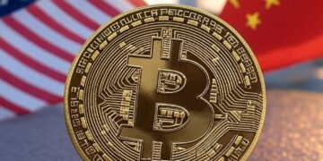 Una moneda dorada con el logo de bitcoin, delante de las banderas de Estados Unidos y China.