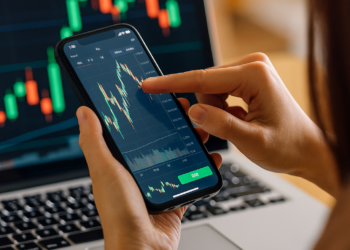 Una persona usando un exchange desde el celular frente a una laptop con gráficos financieros.