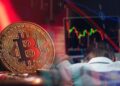 moneda de bitcoin en rojo junto a trader arrepentido frente a computadora con grafico bajista de precio en pantalla y velas de precio superpuestas.