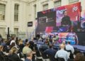 Una vista de un escenario durante un evento llamado "MERGE MADRID", con un gran panel de video mostrando una presentación. En el escenario, cuatro personas están sentadas en sillones, conversando, con una mesa baja en el centro. Delante del escenario, hay una audiencia sentada en sillas negras, con sus espaldas hacia el espectador. El fondo presenta una arquitectura clásica con grandes ventanas.