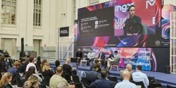 Una vista de un escenario durante un evento llamado "MERGE MADRID", con un gran panel de video mostrando una presentación. En el escenario, cuatro personas están sentadas en sillones, conversando, con una mesa baja en el centro. Delante del escenario, hay una audiencia sentada en sillas negras, con sus espaldas hacia el espectador. El fondo presenta una arquitectura clásica con grandes ventanas.