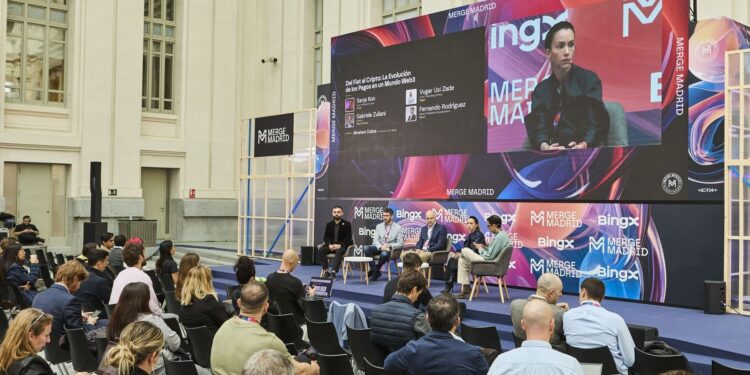 Una vista de un escenario durante un evento llamado "MERGE MADRID", con un gran panel de video mostrando una presentación. En el escenario, cuatro personas están sentadas en sillones, conversando, con una mesa baja en el centro. Delante del escenario, hay una audiencia sentada en sillas negras, con sus espaldas hacia el espectador. El fondo presenta una arquitectura clásica con grandes ventanas.