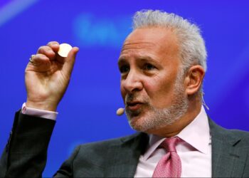 Peter Schiff aparace dando una conferencia y sostiene con su mano derecha una moneda física.