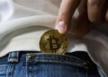 persona sujetando con los dedos moneda de bitcoin mientras la inserta en bolsillo pequeño de pantalon de jean
