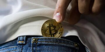 persona sujetando con los dedos moneda de bitcoin mientras la inserta en bolsillo pequeño de pantalon de jean