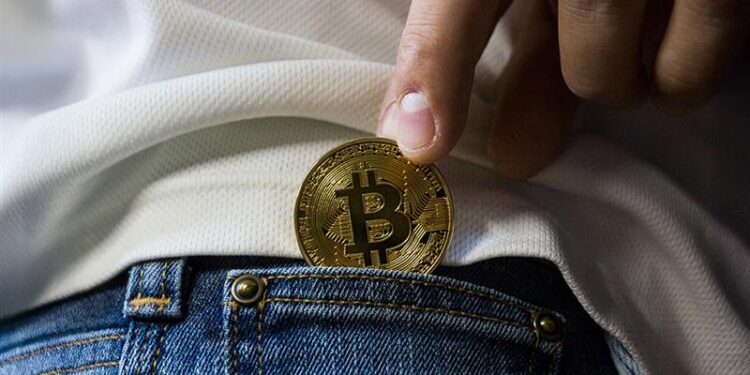 persona sujetando con los dedos moneda de bitcoin mientras la inserta en bolsillo pequeño de pantalon de jean