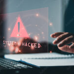 las manos de una persona actuando sobre un techado y un libro abierto mientras se observa un holograma en rojo con la palabra "system hacked"