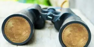 Primer de unos binoculares. A través de las lentes de cristal se observa el reflejo nítido de dos monedas de bitcoin, simbolizando la vigilancia financiera y el monitoreo de su cadena de bloques.