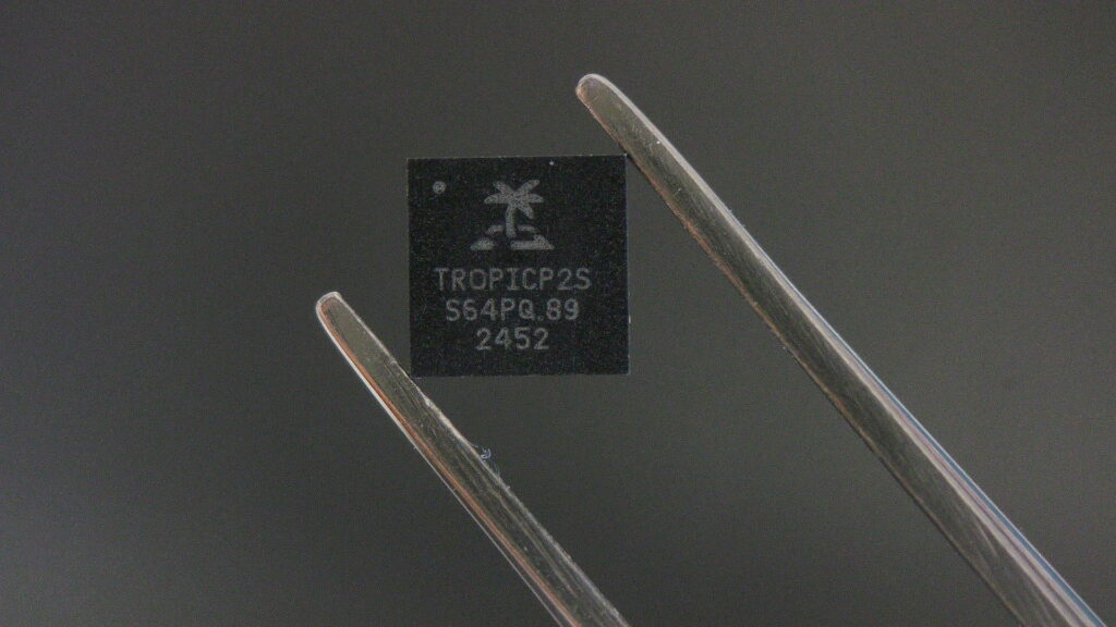 Un chip de color negro con la papalabra tropic sostenido por una pinza plateada, un fondo gris oscuro.