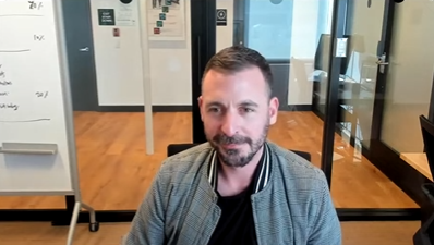 Un hombre con un abrigo gris y una remera negra sentado en una silla en una videoconferencia.
