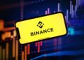 Logo de Binance sobre una pantalla de un móvil con velas rojas y verdes de fondo que reflejan la volatilidad del mercado.
