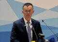 Changpeng Zhao, fundador de Binance, habla frente a un micrófono en un acto oficial en Kirguistán, vestido con traje oscuro y corbata, con un fondo azul geométrico.