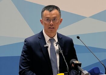 Changpeng Zhao, fundador de Binance, habla frente a un micrófono en un acto oficial en Kirguistán, vestido con traje oscuro y corbata, con un fondo azul geométrico.