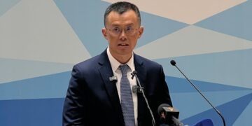 Changpeng Zhao, fundador de Binance, habla frente a un micrófono en un acto oficial en Kirguistán, vestido con traje oscuro y corbata, con un fondo azul geométrico.