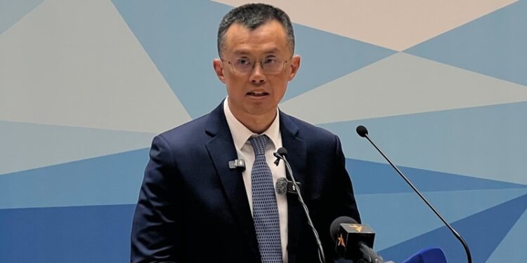 Changpeng Zhao, fundador de Binance, habla frente a un micrófono en un acto oficial en Kirguistán, vestido con traje oscuro y corbata, con un fondo azul geométrico.