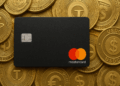 Tarjeta negra de Mastercard apoyada sobre un conjunto de monedas doradas que representan las stablecoins USDT y USDC, con sus logotipos grabados en relieve.
