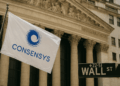 Bandera blanca con el logotipo azul de ConsenSys ondeando frente a la fachada del edificio de la Bolsa de Nueva York en Wall Street, junto al letrero negro característico de “WALL ST”.