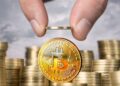 monedas físicas de bitcoin doradas apiladas una encima de otra, mientras dos dedos sostienen otra moneda de bitcoin.