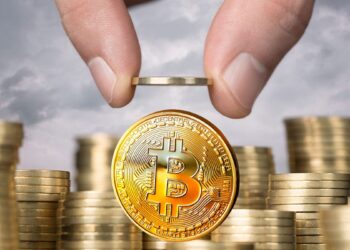 monedas físicas de bitcoin doradas apiladas una encima de otra, mientras dos dedos sostienen otra moneda de bitcoin.